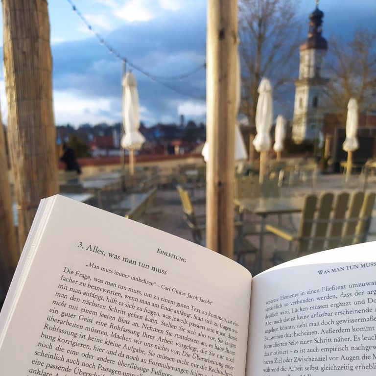 Aufgeschlagenes Buch im Vordergrund. Unscharf im Hintergrund der Blick vom Freisinger Domberg auf die Kirche St. Georg und die Altstadt.