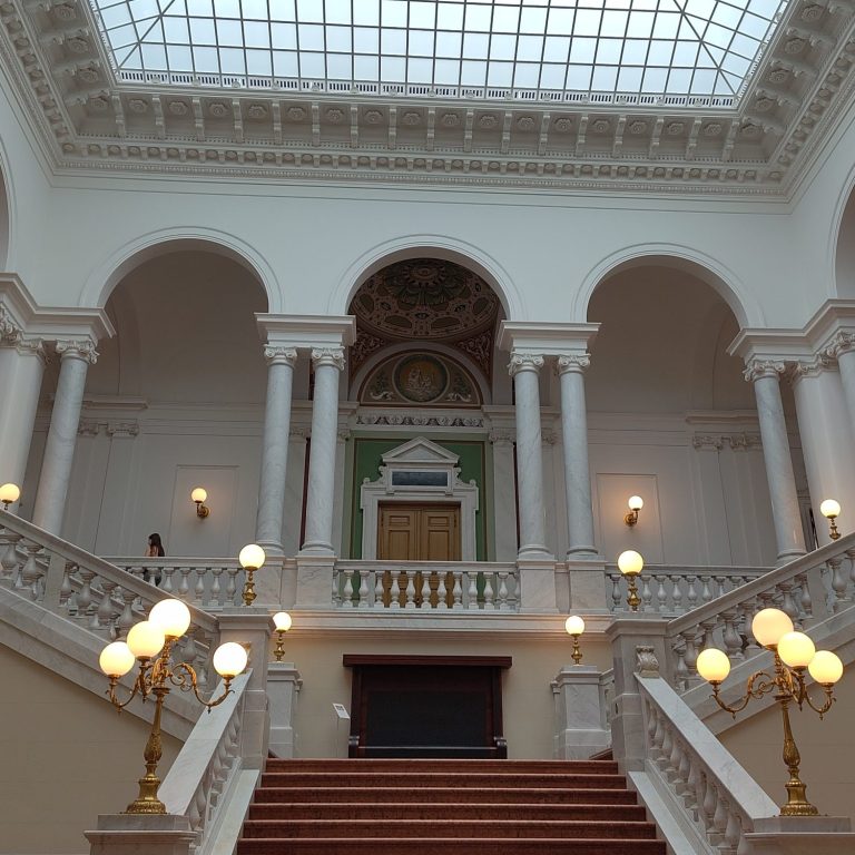 Ein Foto von Treppenaufgang in der Leipziger Bibliothek für Geisteswissenschaften. Rote Stufen mit goldenen Leuchten und weißem Geländer.