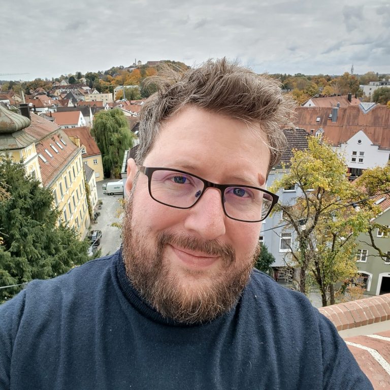 Selfie von Max Breu in dunkelblauem Pullover von einer erhöhten Plattform mit Blick über die Freisinger Altstadt mit roten Ziegeldächern.